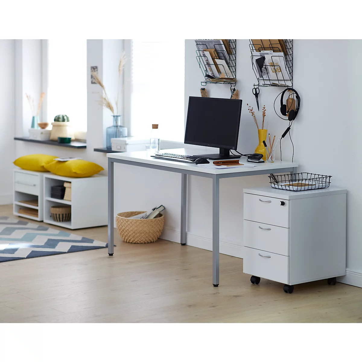 Een minimalistische werkplek met bureau, computer, opbergruimte en ladekast. Gele kussens op een kleine bank bij het raam.