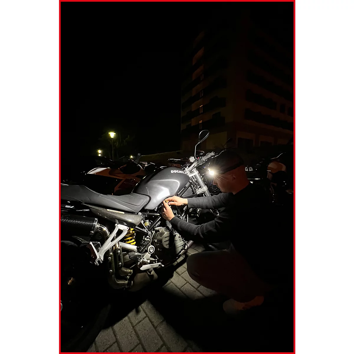 Mann repariert Motorrad im Dunkeln. Im Hintergrund ein Wohnhaus und weitere Fahrzeuge.