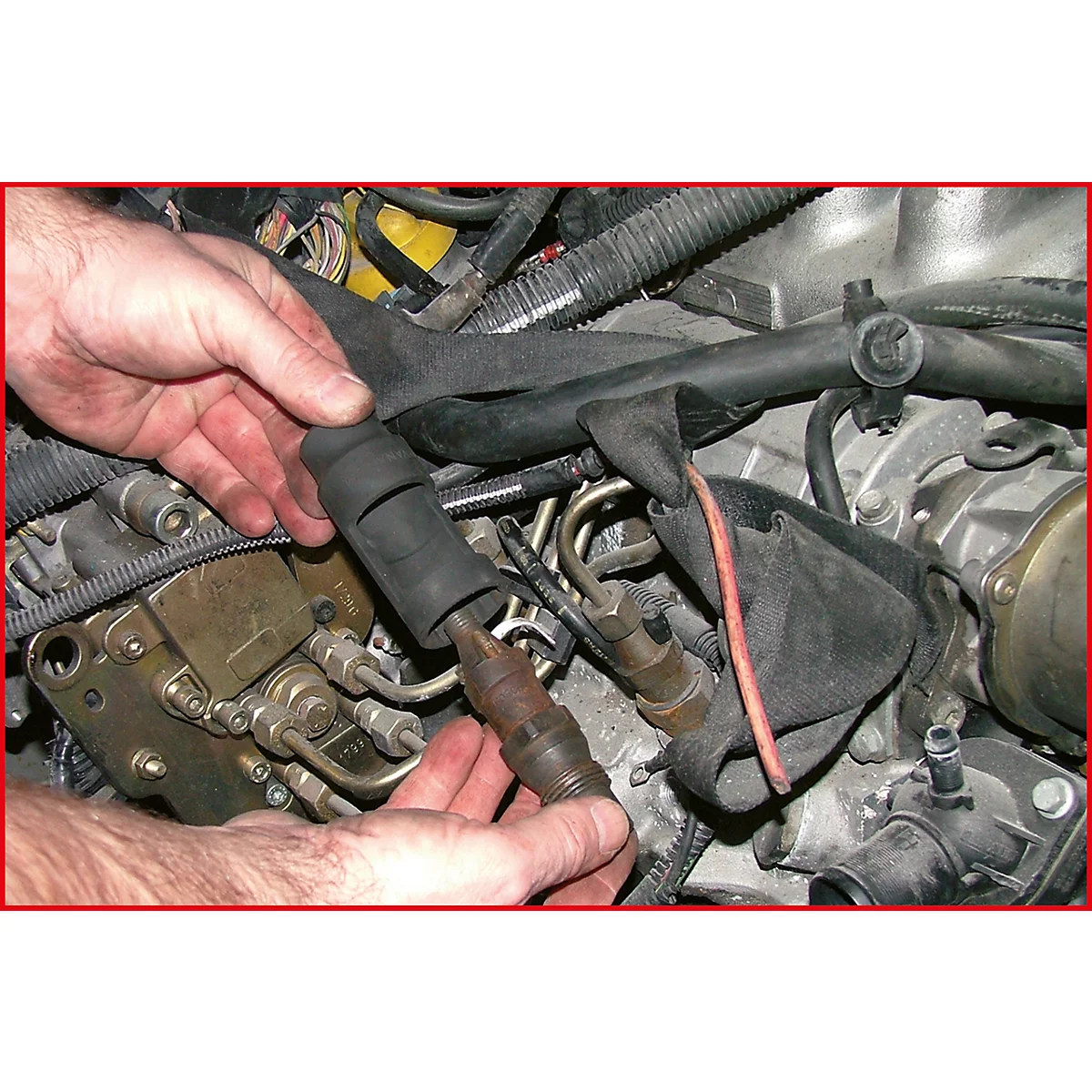 Nahaufnahme der Hände, die einen defekten Zündstecker aus einem Motor ausbauen. Sichtbar sind Schläuche, Kabel und weitere Motorteile.