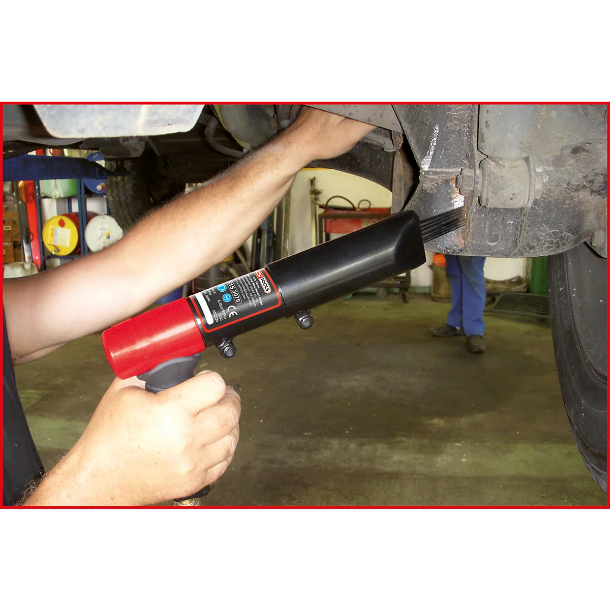 Mechaniker arbeitet mit einem Druckluftnagler an einem Auto. Der Nagler ist schwarz und rot. Im Hintergrund ein weiterer Mechaniker.