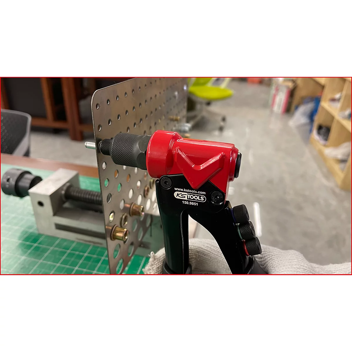 Nahaufnahme eines roten Nietwerkzeugs, das auf einer Metallplatte befestigt ist, auf der sich mehrere Nieten befinden, wobei eine Schraubzwinge im Hintergrund zu sehen ist.