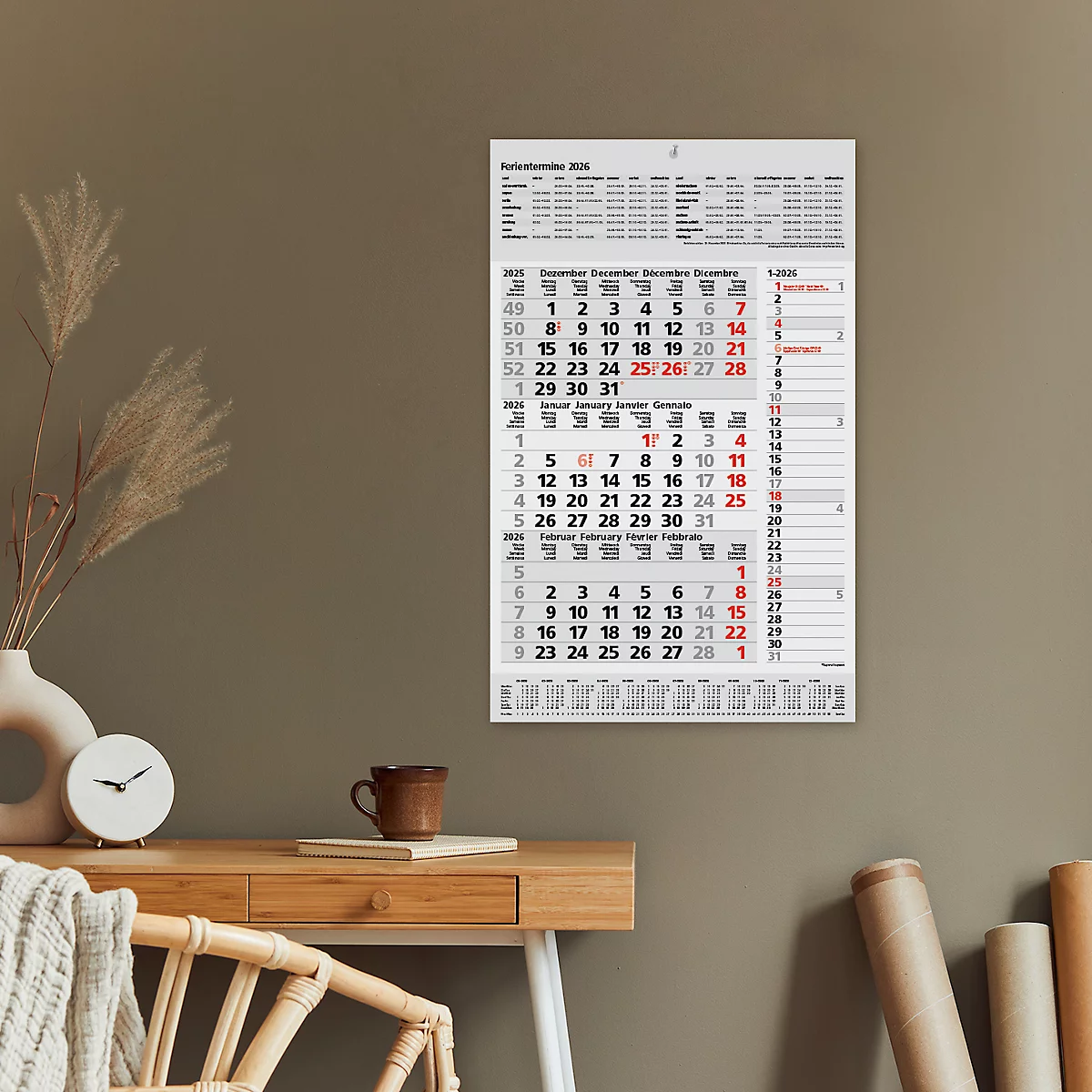 Wandkalender mit Monatsübersicht hängt an einer Wand über einem Schreibtisch. Auf dem Schreibtisch liegen ein Becher, eine Uhr und ein dekorativer Überwurf.