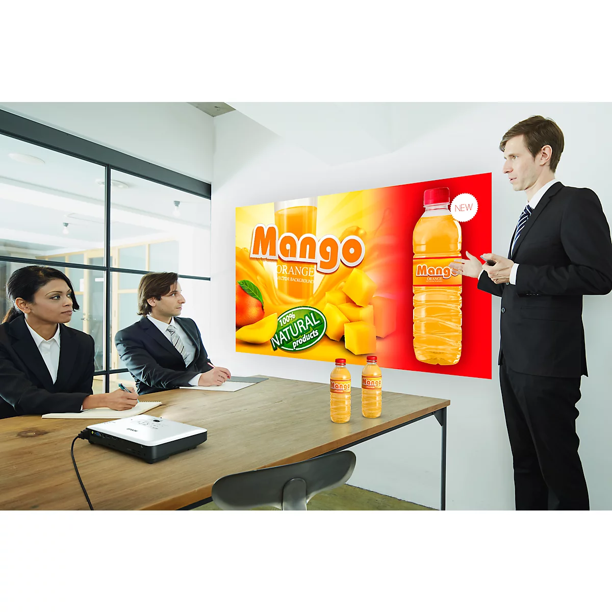 A man presents a Mango drink in a presentation. Two listeners. Projection, bottles on table.