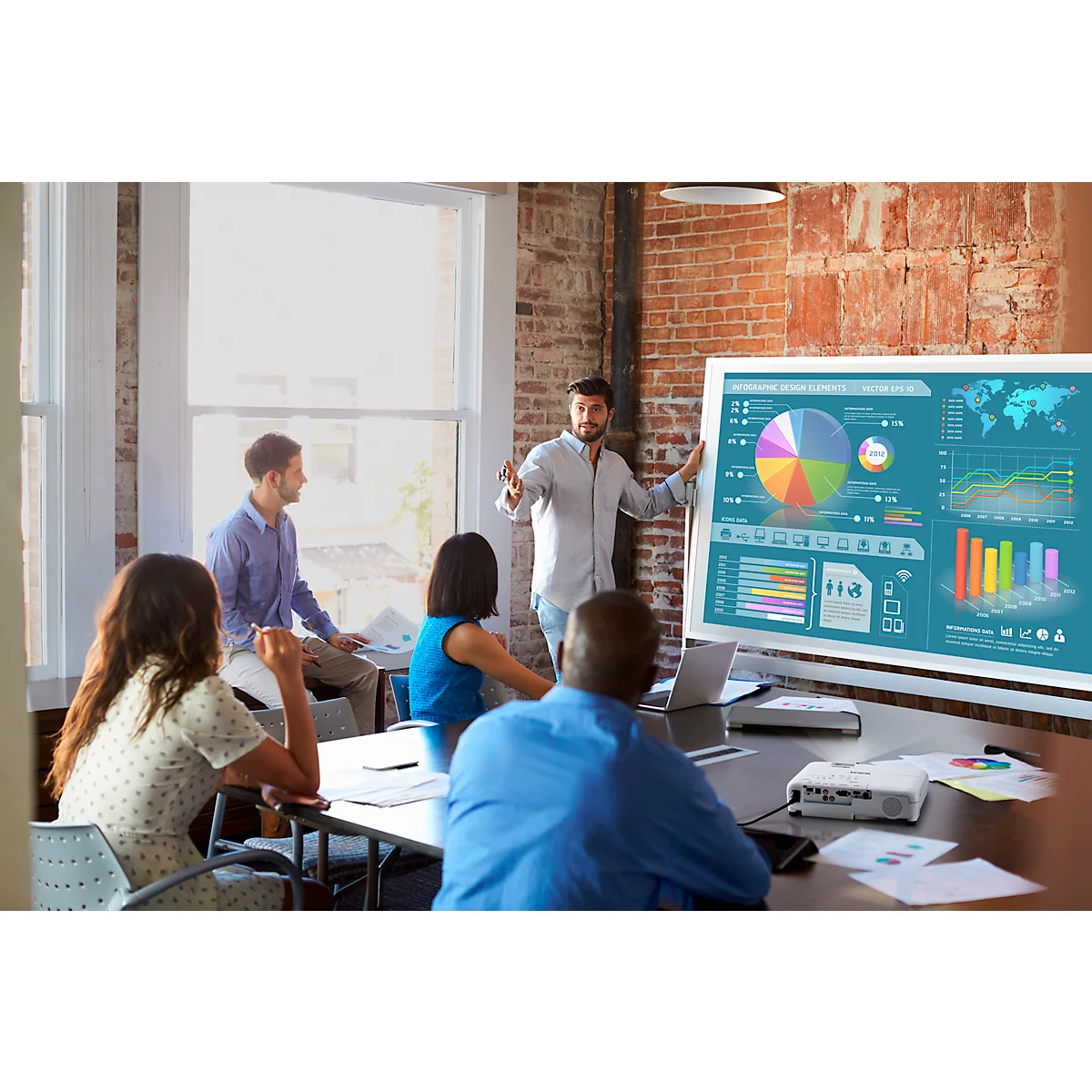 A presentation in a conference room. A man stands in front of a screen with diagrams. Other people sit at the table.