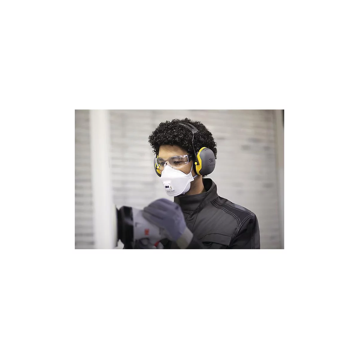 Een man met krullend haar draagt een veiligheidsbril, een masker, oorkappen en handschoenen. Hij schuurt iets met een machine.