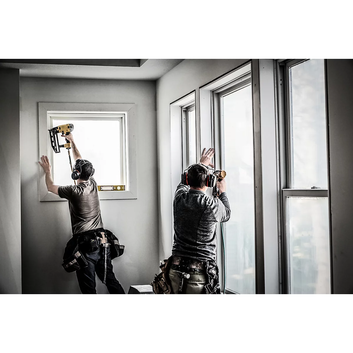 Zwei Handwerker installieren Fenster. Einer nagelt, der andere schraubt. Beide tragen Gehörschutz und Werkzeugtaschen.