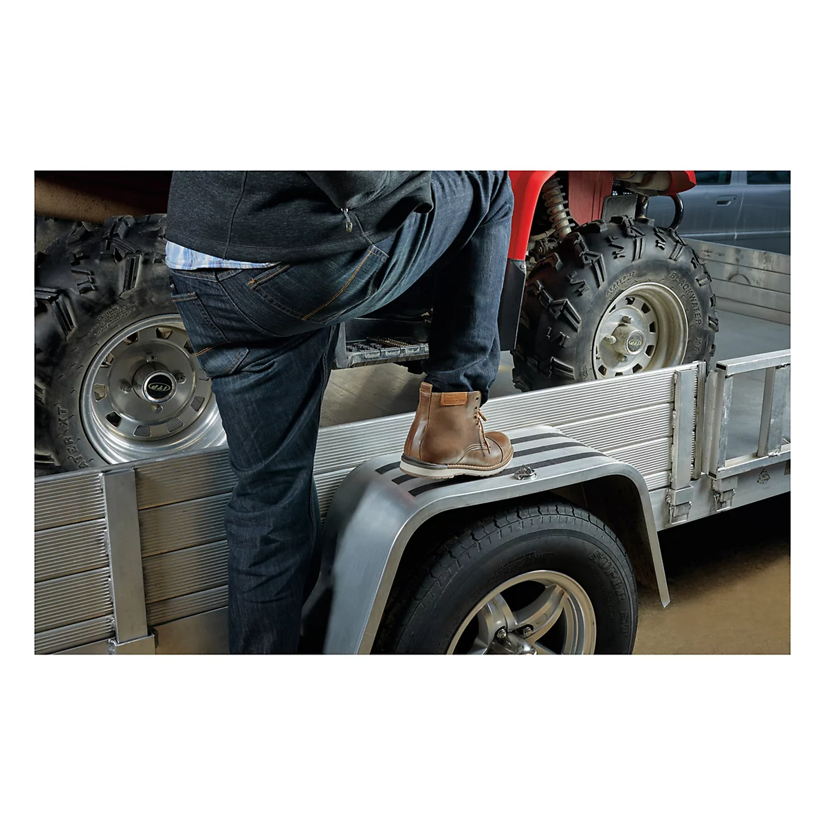 Eine Person steigt auf einen Anhänger. Das rechte Bein steht auf dem Kotflügel, das linke Bein auf der Ladefläche. Im Hintergrund ein rotes ATV.