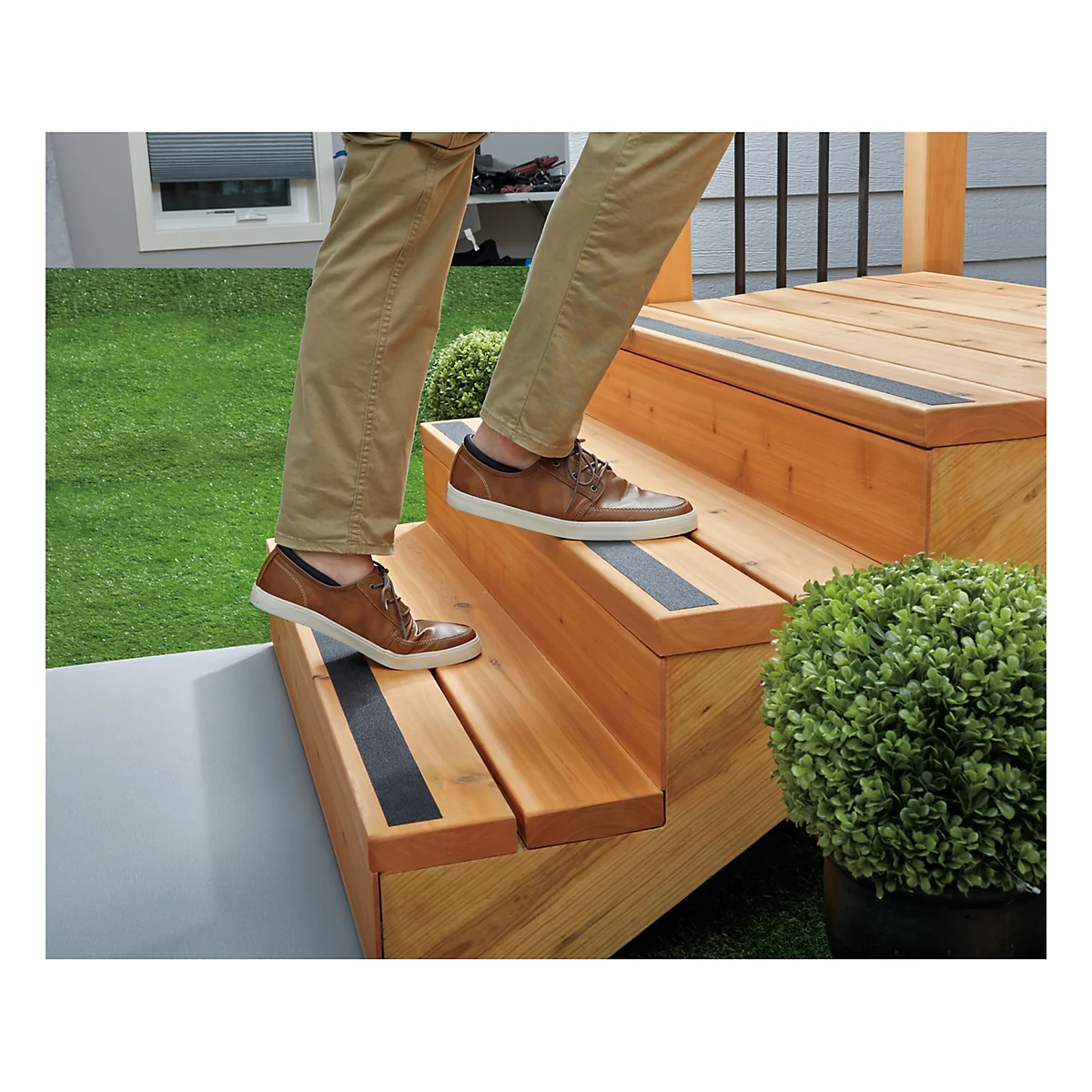 Person steigt Holztreppe hinauf. Auf jeder Stufe ein schwarzer Streifen. Auf einer Seite grüner Rasen, im Hintergrund ein Fenster.