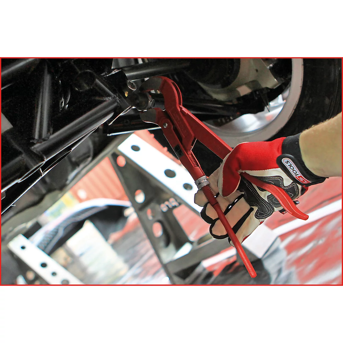 Person in Handschuhen repariert mit roter Zange am Unterboden eines Fahrzeugs. Metallteile, Reifen und Werkzeug sichtbar.