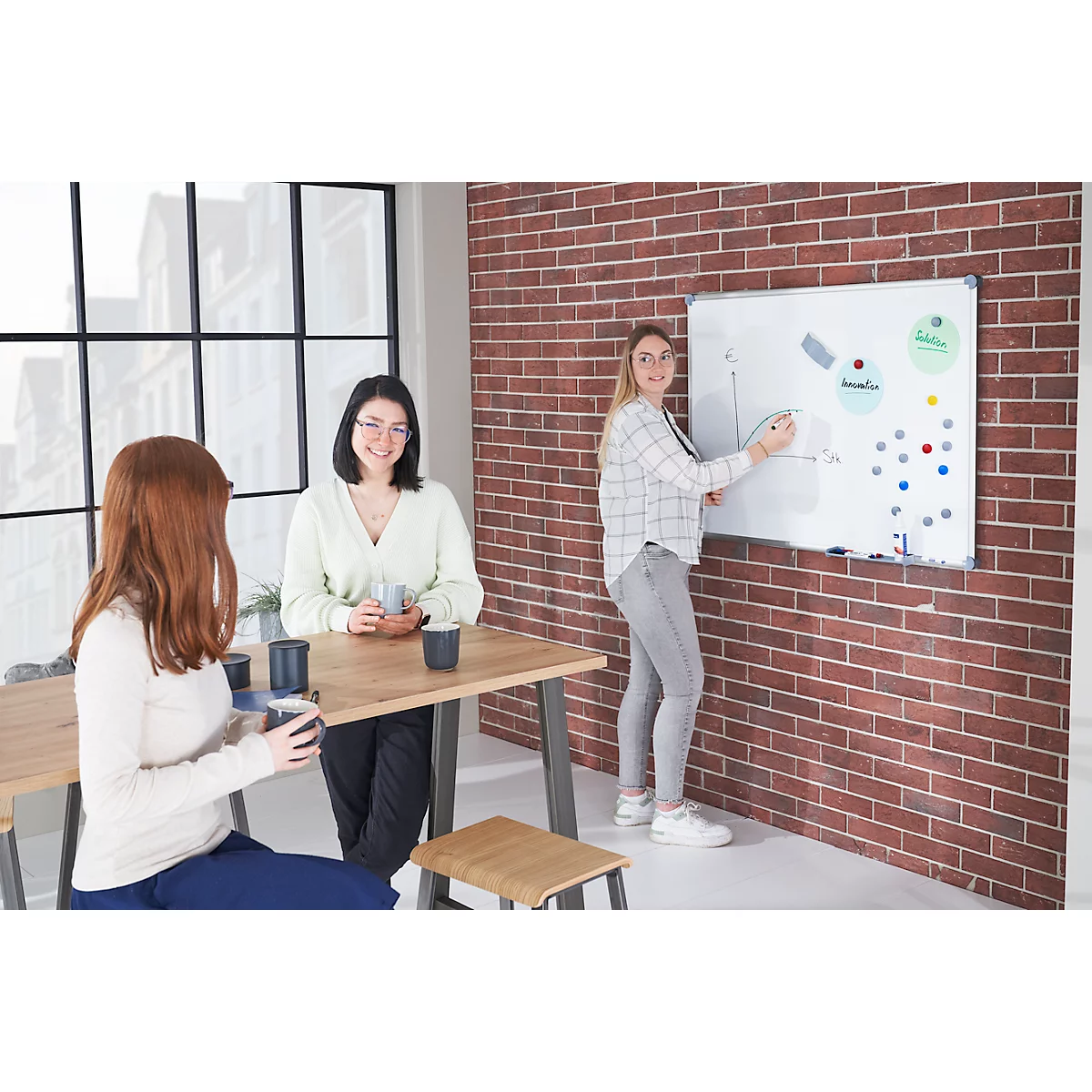 Trois femmes dans un bureau : une explique quelque chose sur le tableau blanc, deux sont assises à la table avec du café.