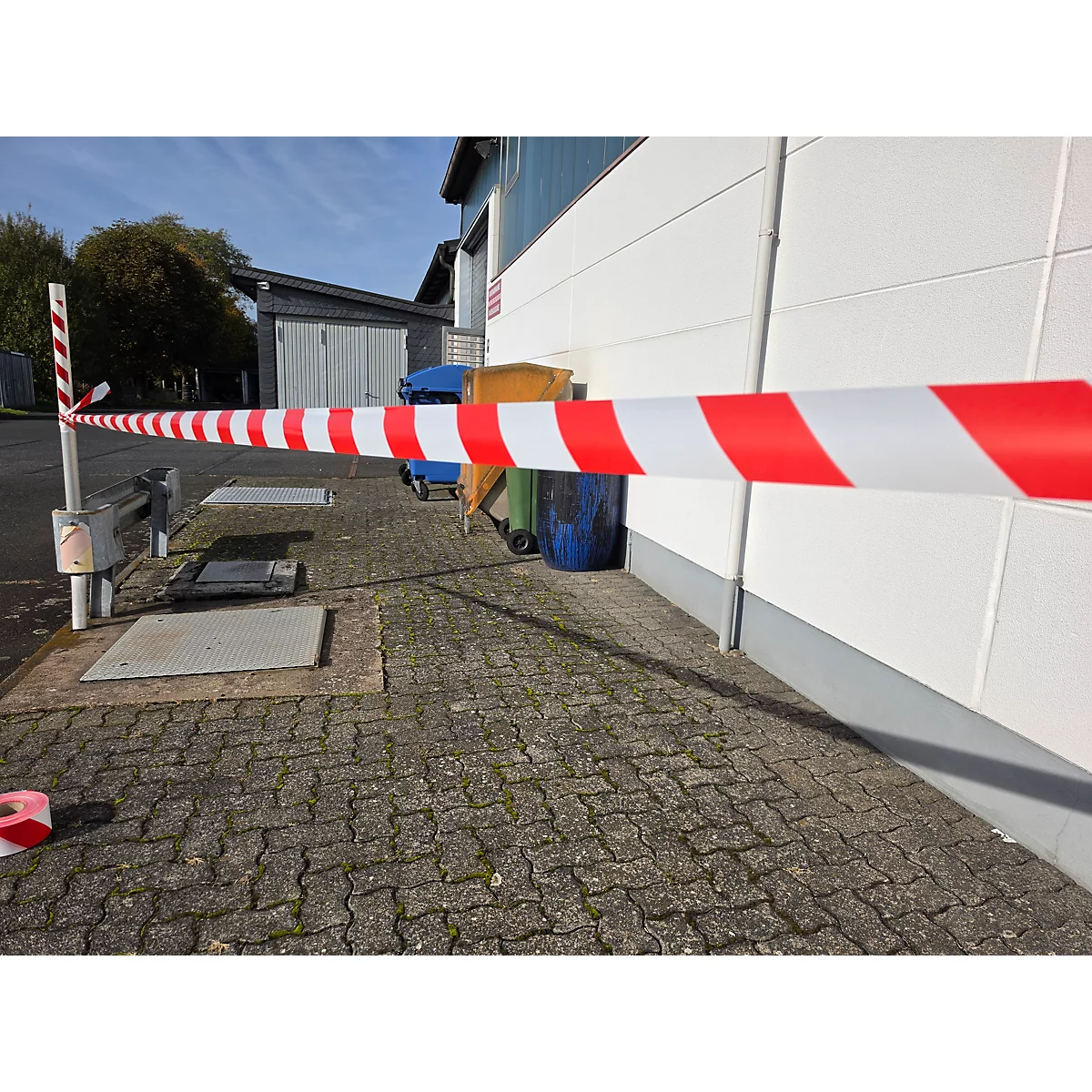 Rot-weißes Absperrband spannt sich vor einer Lagerhalle auf einem gepflasterten Außenbereich.