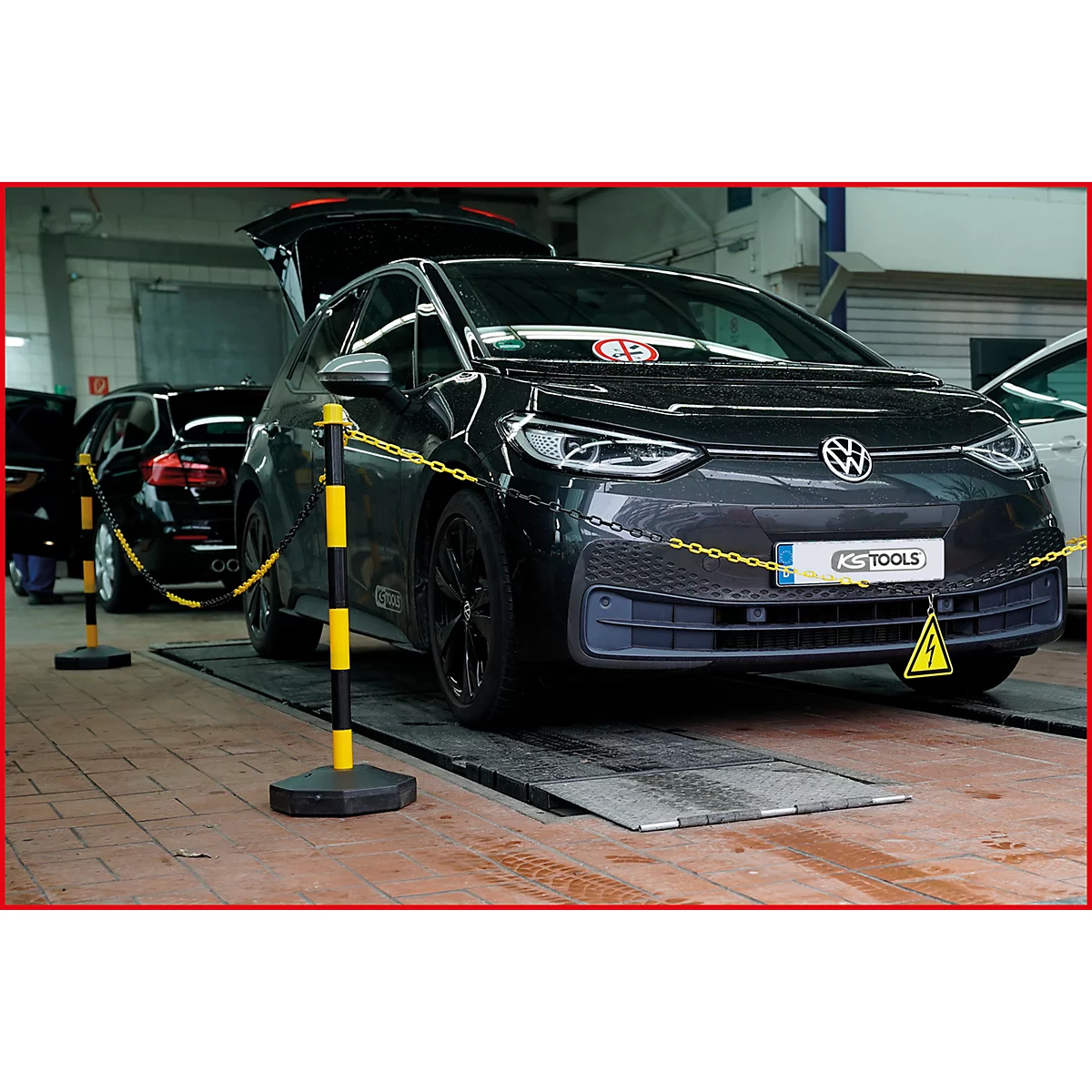 Dunkelgraues VW-Elektroauto in einer Werkstatt, Motorhaube offen. Gelb-schwarze Absperrpfosten mit Kette, Warndreieck.