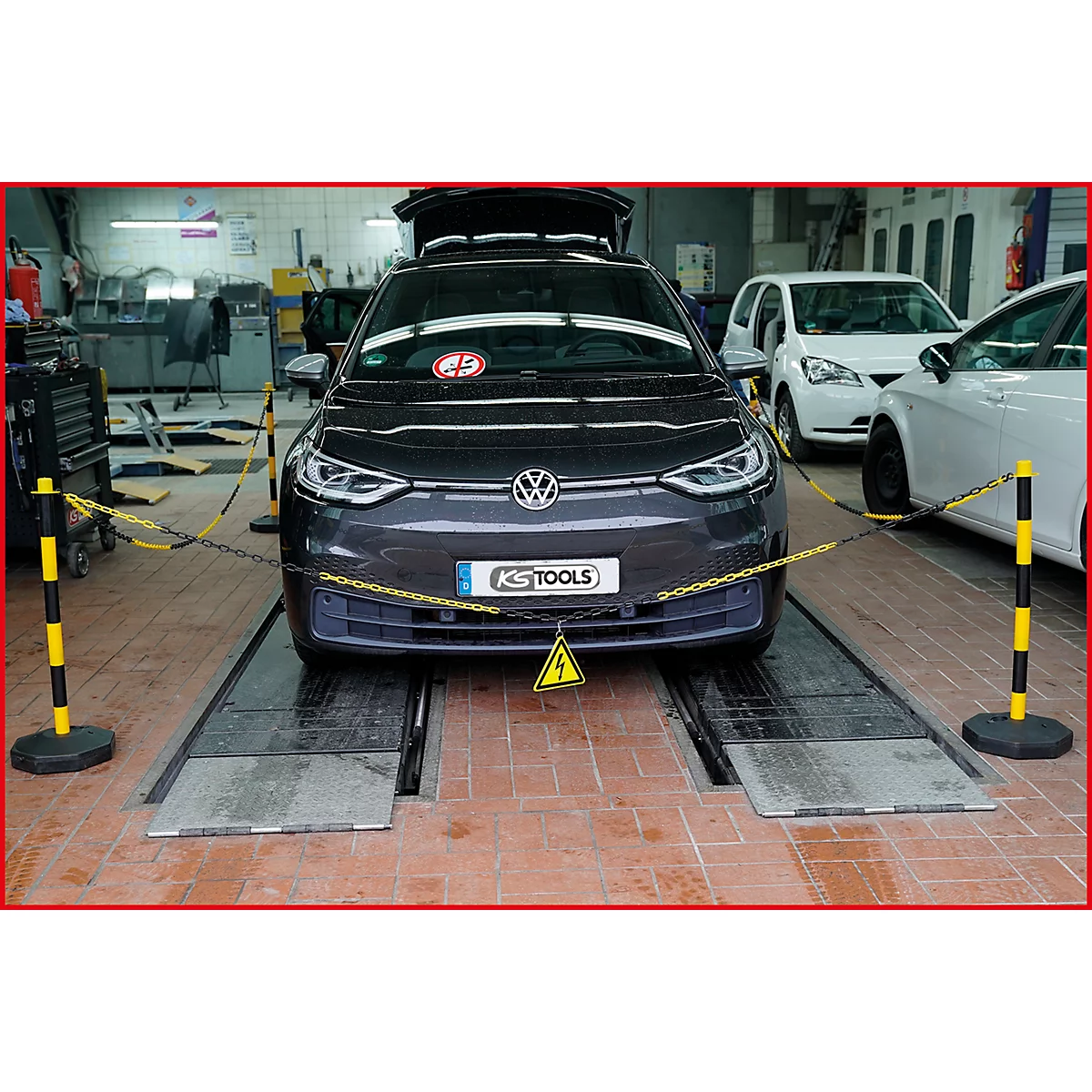 Ein graues VW-Elektroauto steht in einer Werkstatt auf einer Hebebühne, gesichert mit Ketten. Ein Achtung-Symbol ist sichtbar.