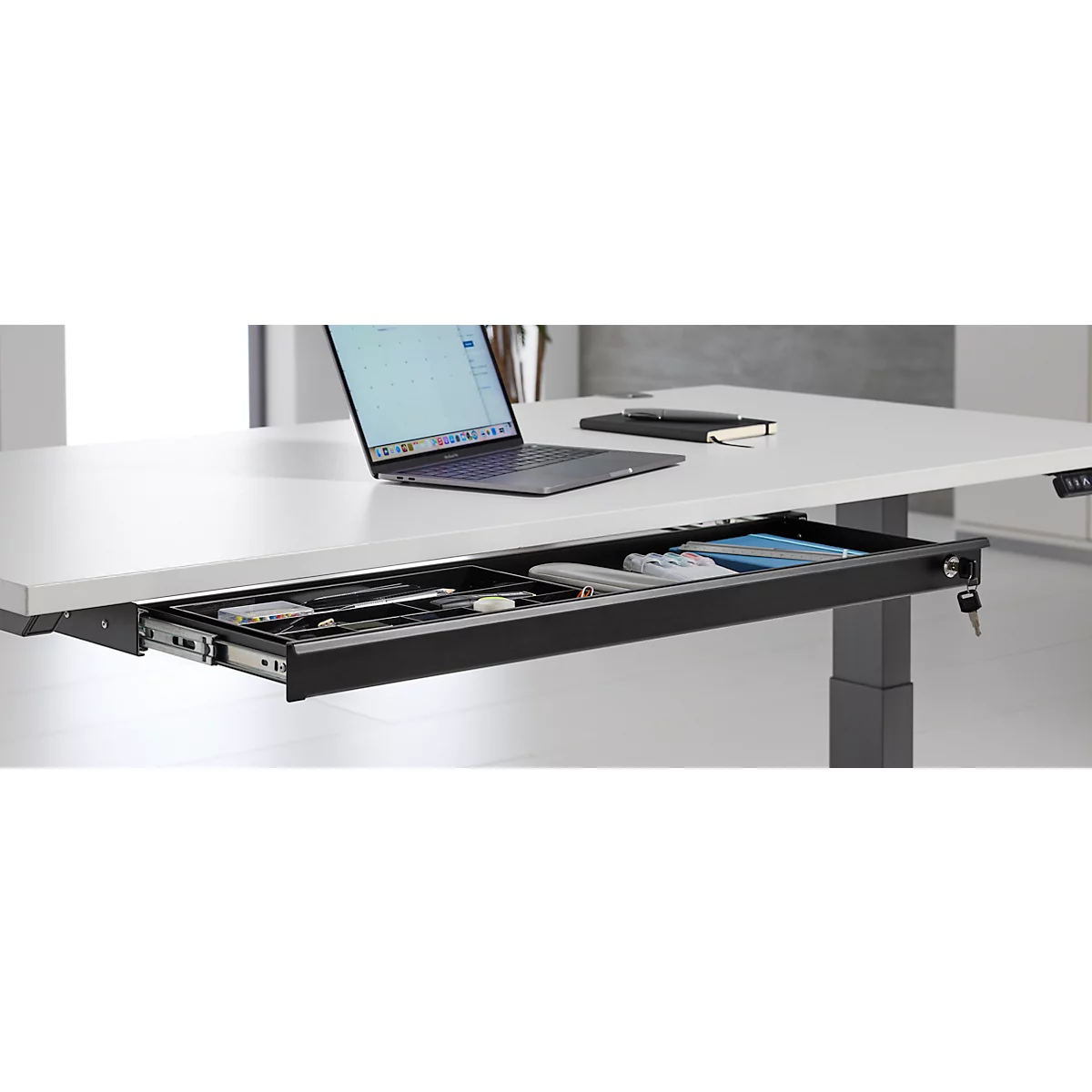 Bureau met uitgetrokken lade. Laptop, notitieboek en pennen liggen op het tafelblad.