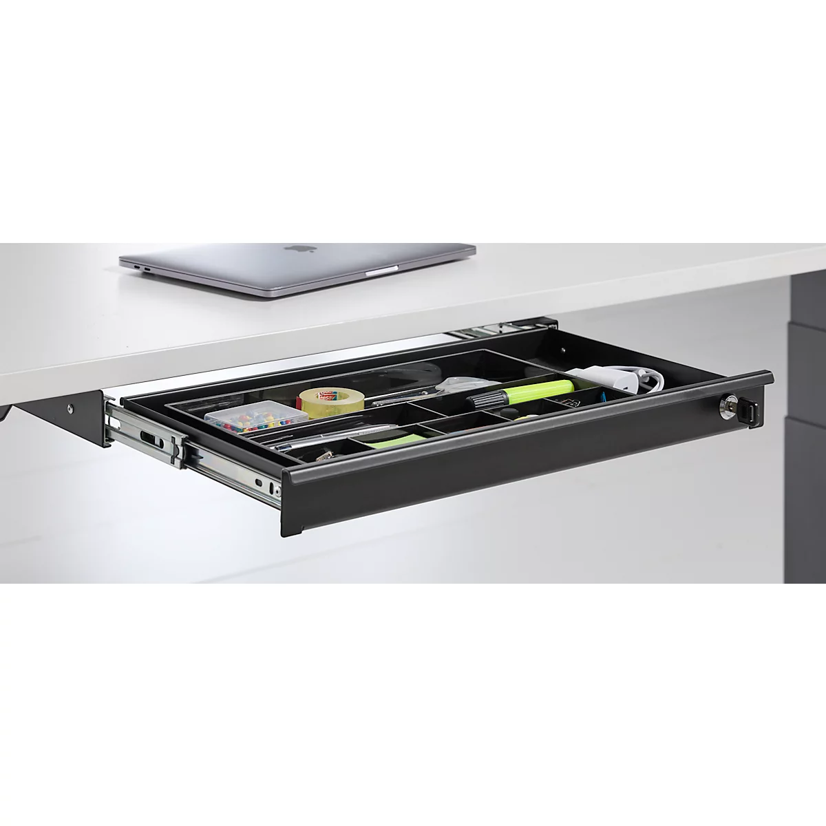 Uitgetrokken lade onder een bureau met pennen, tape, markeerstift en notities. Een laptop staat erboven.