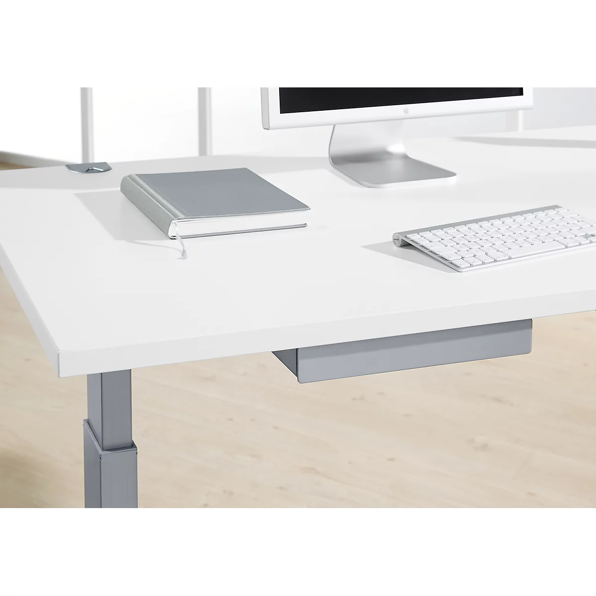 White desk with monitor, keyboard, and notebook. Below, a drawer. The desk stands on a wooden floor.