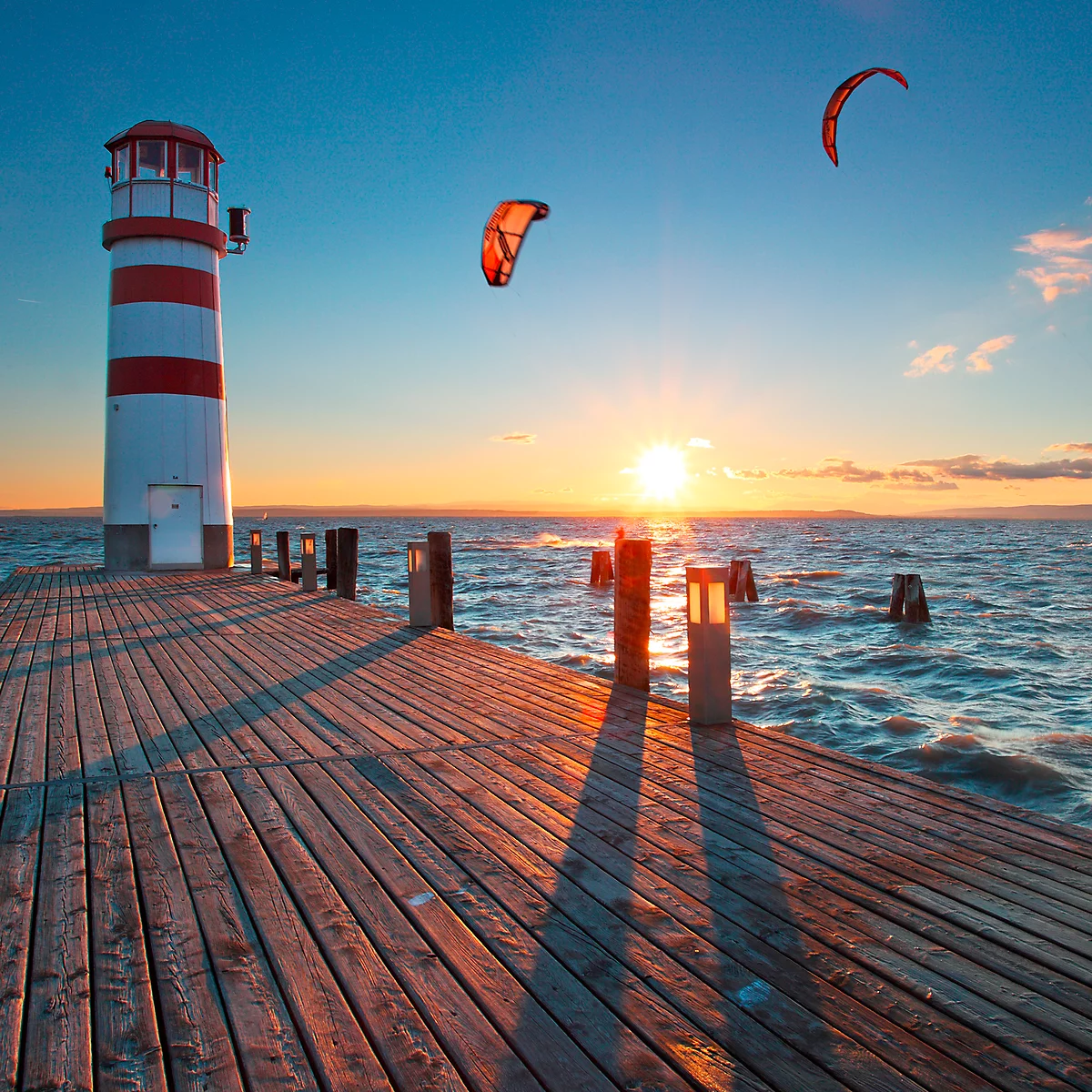 Lighthouse with red and white stripes, pier and kitesurfers at sunset.