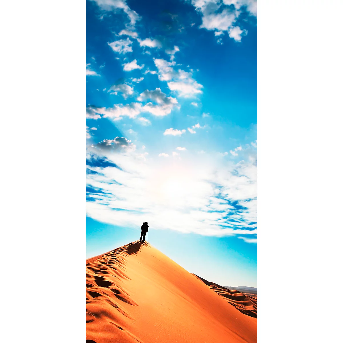 Hiker stands on a sand dune, looking afar, blue sky, sun, clouds.