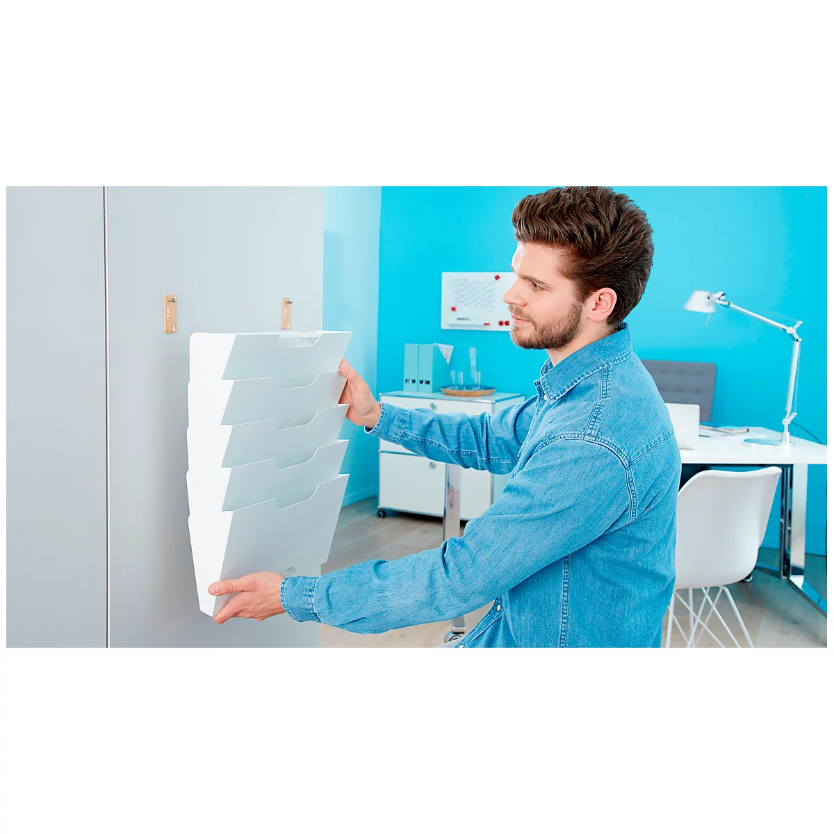 A man attaches a white wall brochure holder. He is in an office with a blue background.