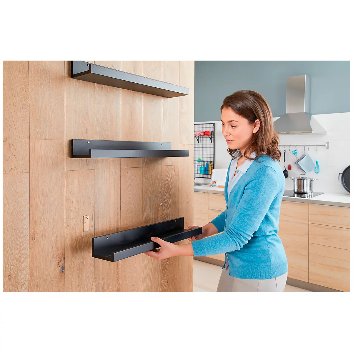 Woman installing a black wall shelf on a wooden wall. The kitchen is visible in the background.