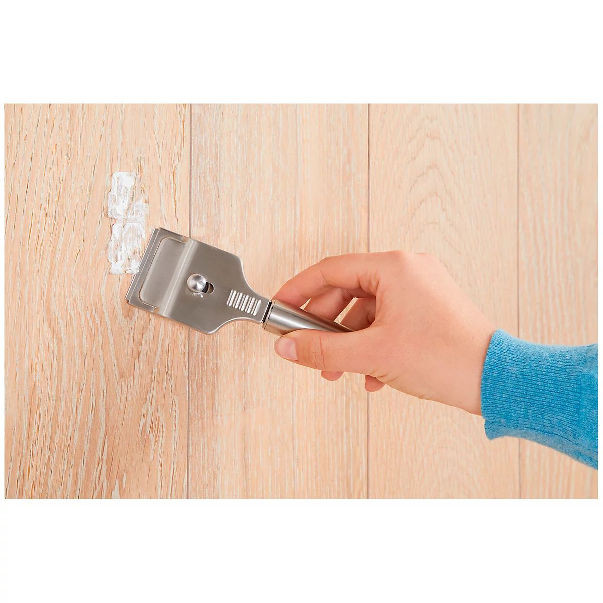 A hand using a metal scraper to remove paint from a wooden surface.