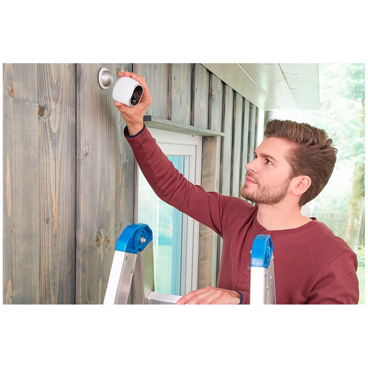 A man installs a security camera on a house wall. He is standing on a ladder and holding the camera towards the bracket.
