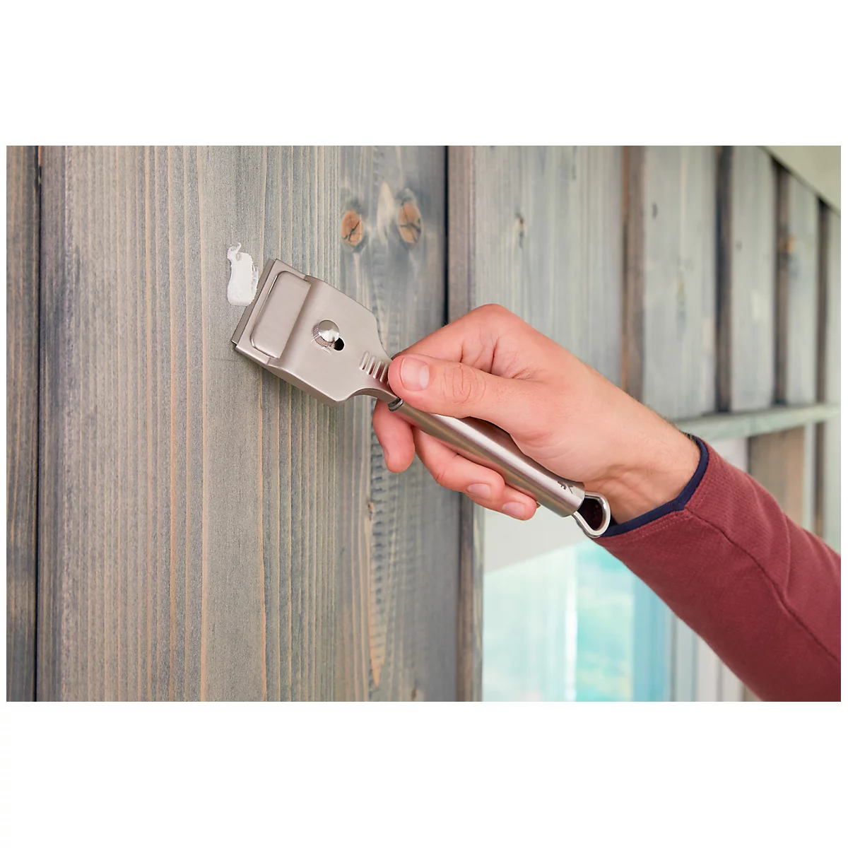 Hand holding scraper on grey wooden surface. Putty is being removed.