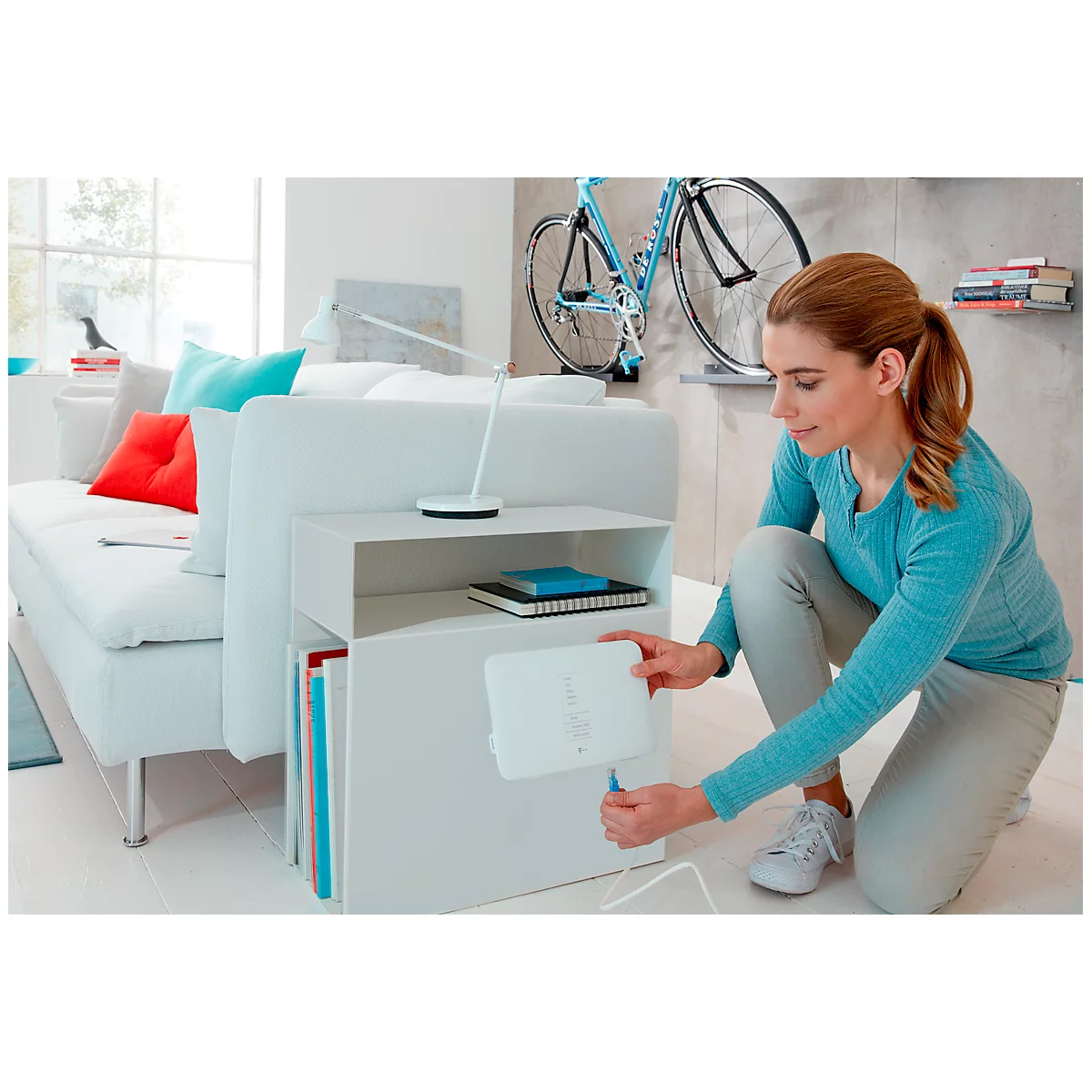 A woman kneeling, plugs a cable into a white box attached to a white side table.
