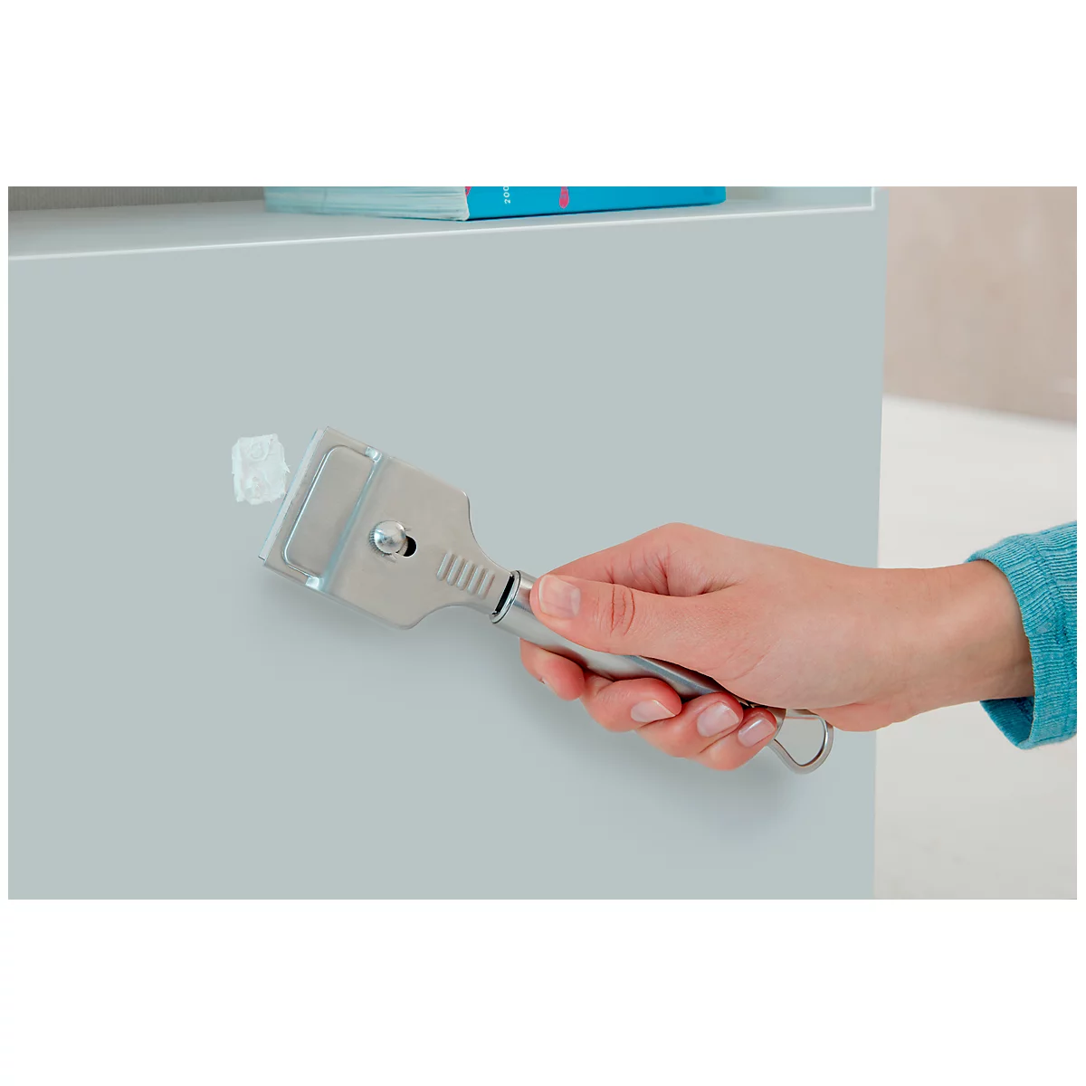 A hand holds a silver scraper against a light gray wall. A piece of white plaster has been removed.