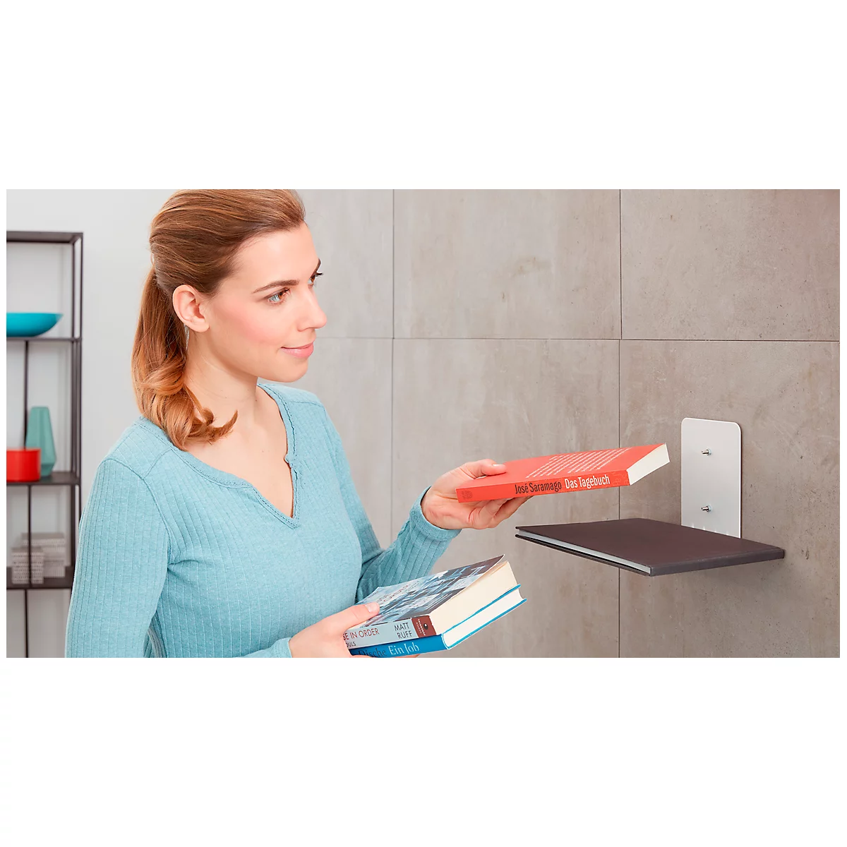 A woman places books on a wall shelf, near a shelf wall.