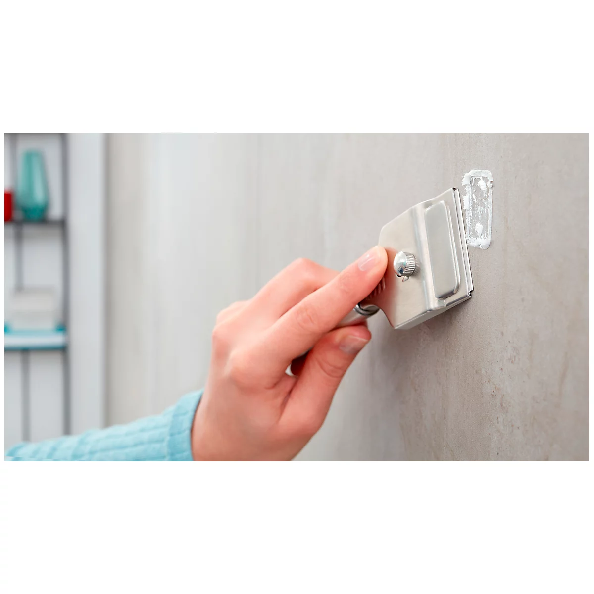 Hand holding a silver blade against the wall. A white adhesive hook is already attached to the wall.