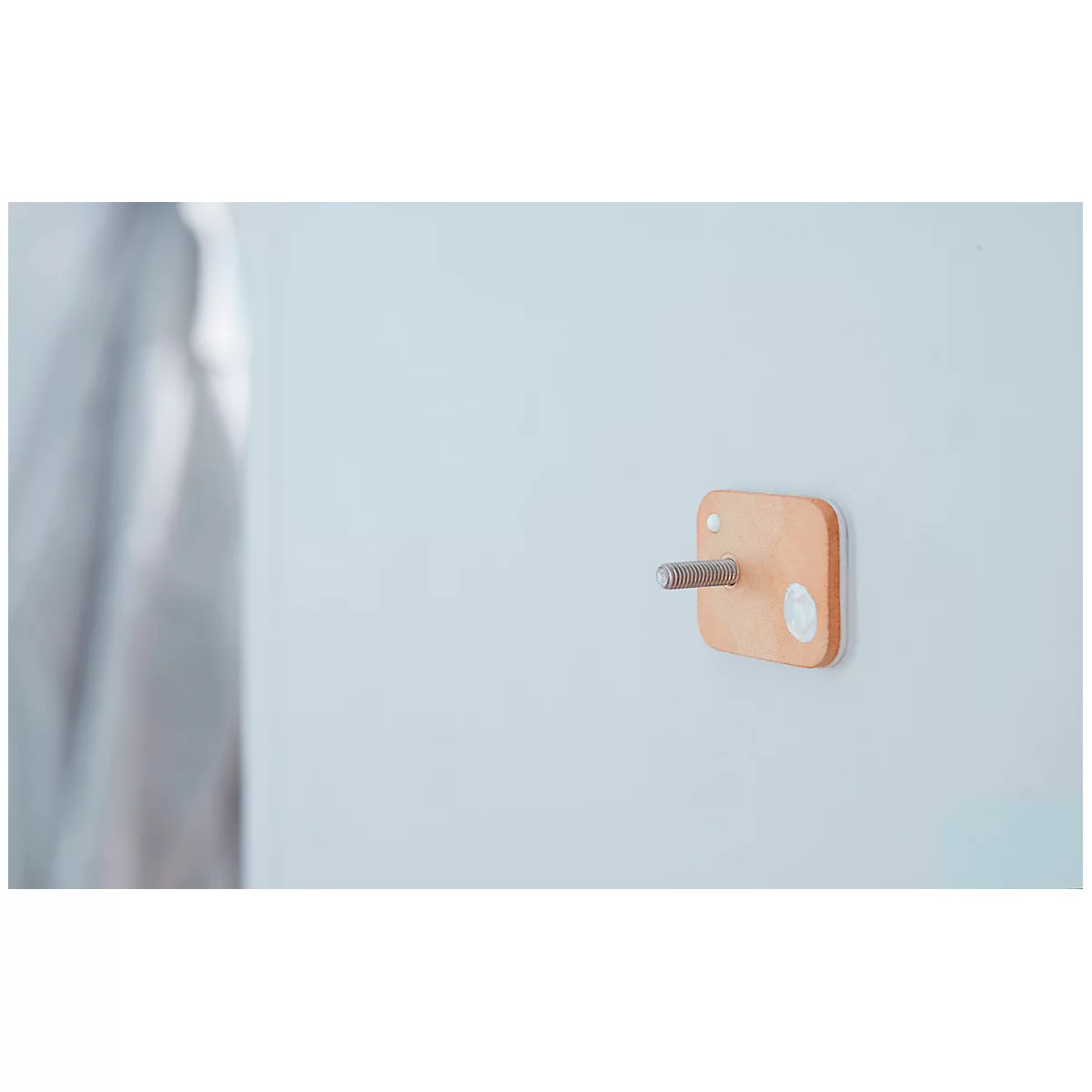 A square, wood-colored wall hook with a silver pin, attached to a white wall. To the right of it is a bright spot.