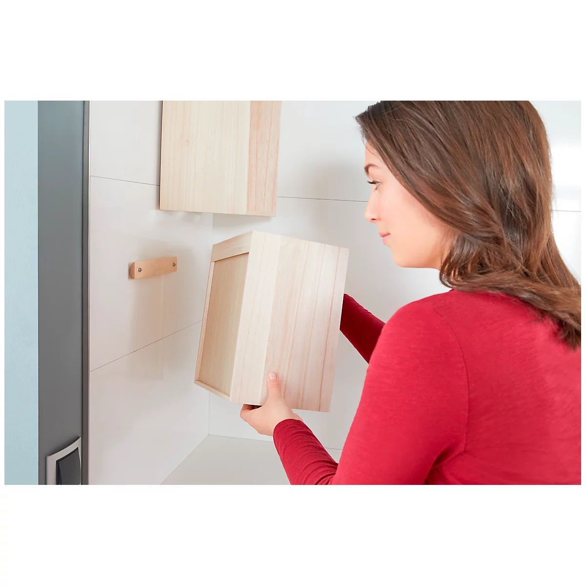 A woman in a red blouse is attaching a wooden box to a wall. Another box and a bracket are also visible.