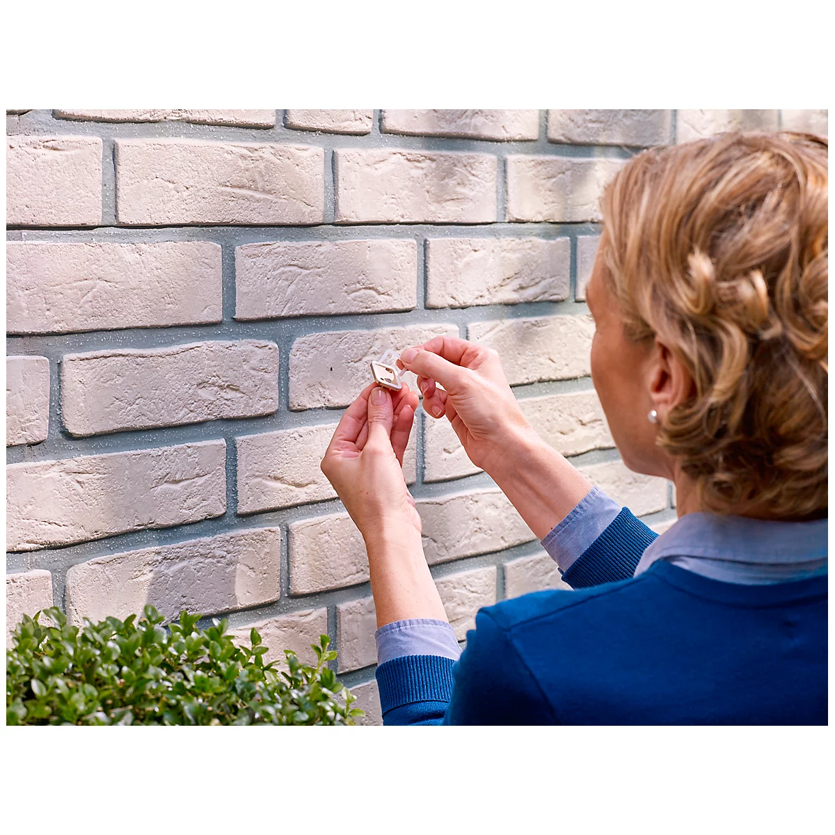 Woman attaching a hook to a brick wall. She is wearing a blue top and has blonde hair.