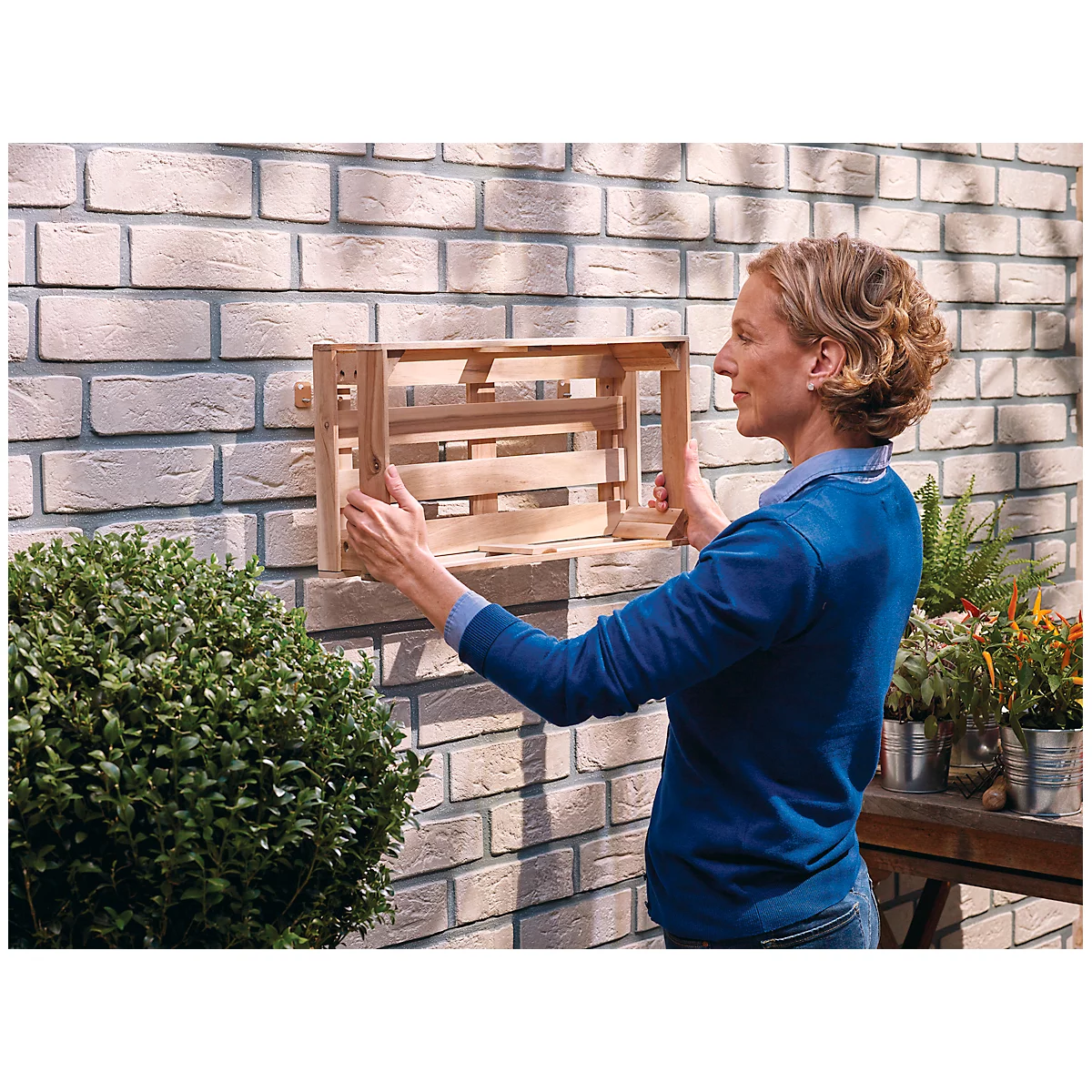 Woman mounting wooden crate on brick wall. Plants in buckets next to it.
