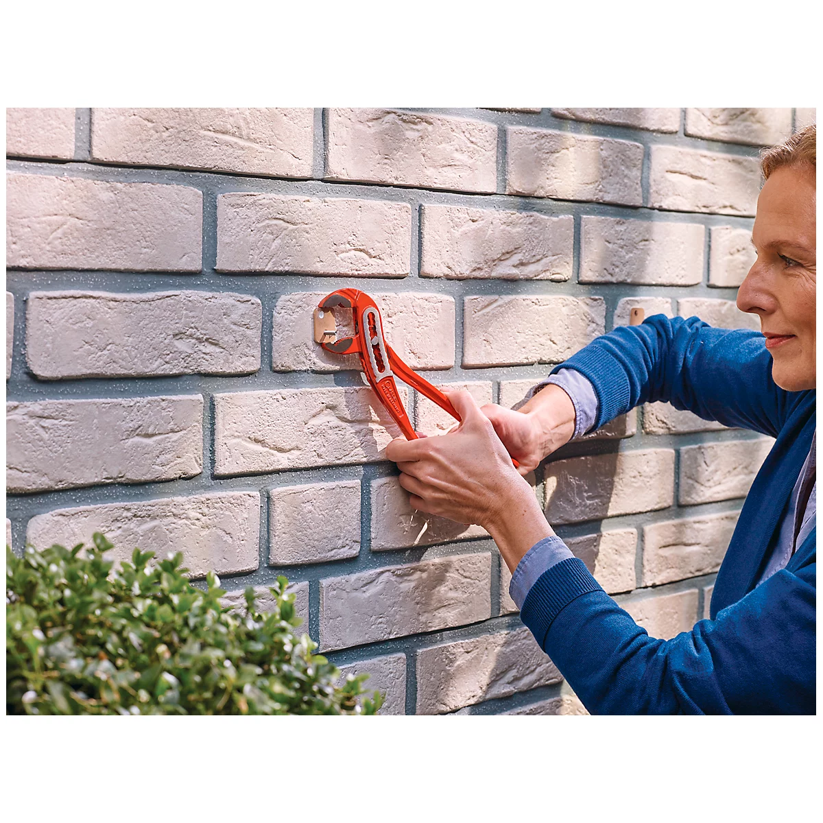A woman is mounting something on a brick wall with red pliers.