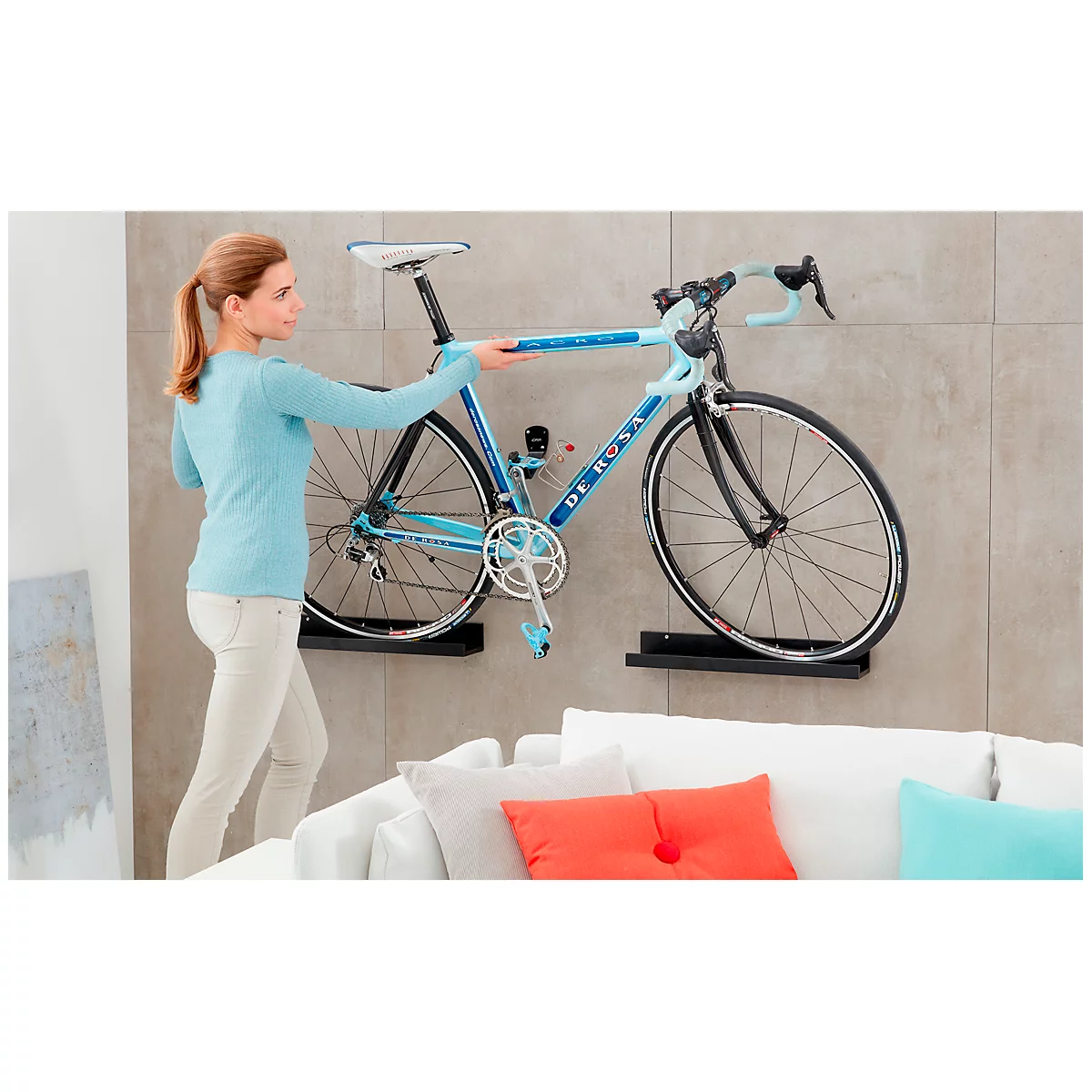 Woman hanging a bicycle on a wall mount. Light blue bike with 'De Rosa' lettering, gray wall, white sofa.