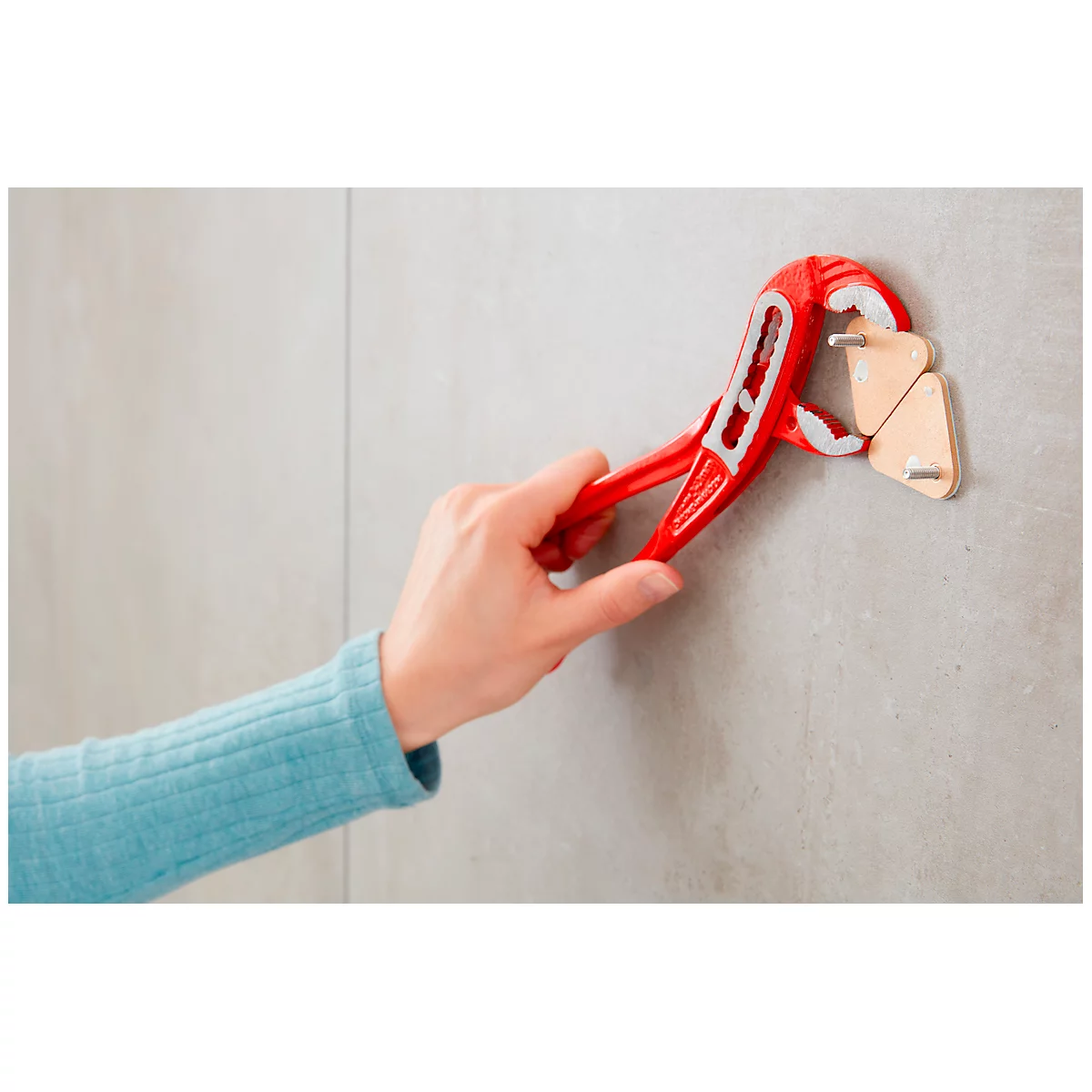 Hand holding red pliers attaching two triangular wooden pieces to a wall.