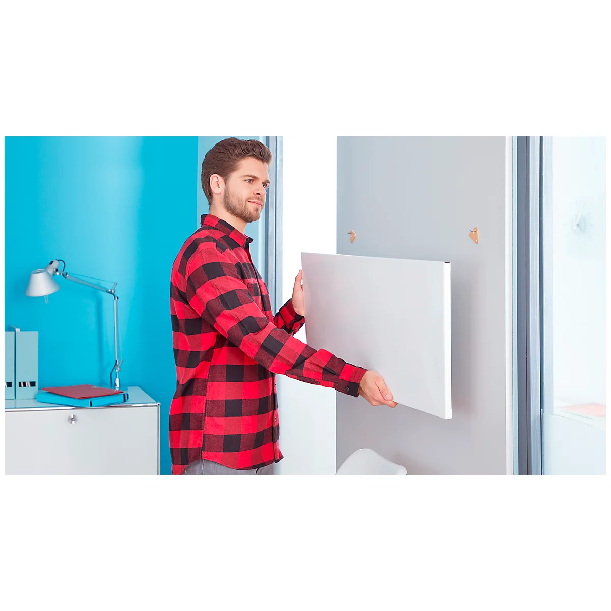 A man in a red and black checkered shirt hangs a white picture.