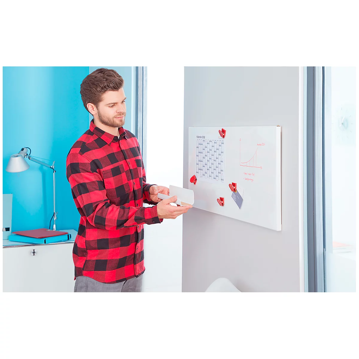 A man in a red plaid shirt stands in front of a magnetic whiteboard. He is looking at a white sheet of paper in his hands.