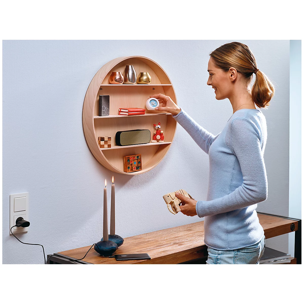 A woman places an alarm clock on a round wooden shelf. A speaker, books, and toys are on the lower shelves. Two candles are burning below.