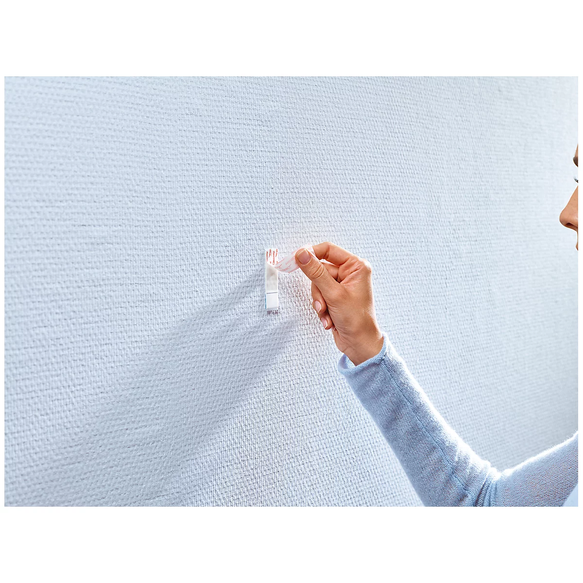 A person holds a sticky hook to a textured white wall. The person's face is partially visible.