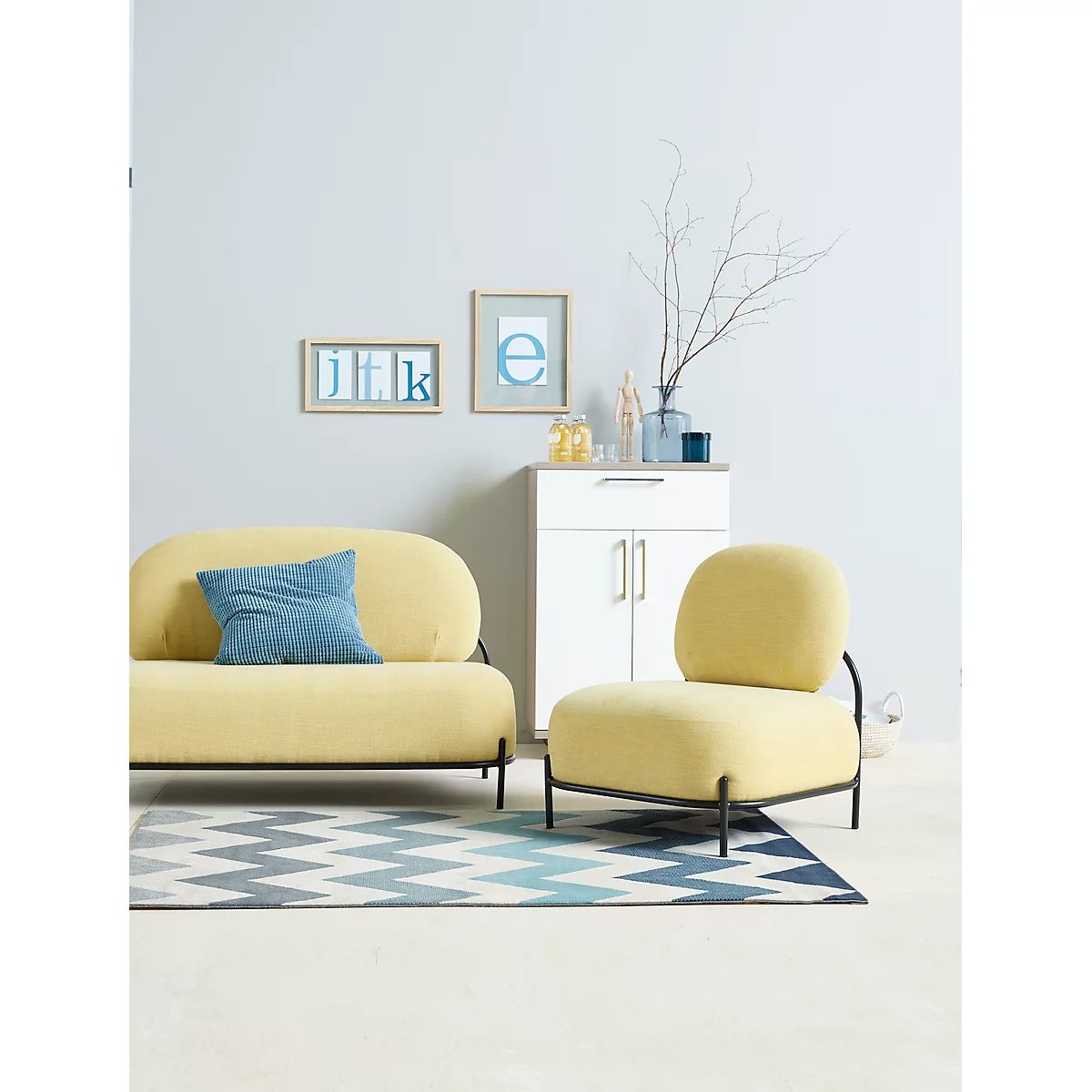 Yellow sofa and armchair with black metal frames on a rug. In the background a cabinet with decoration and pictures.