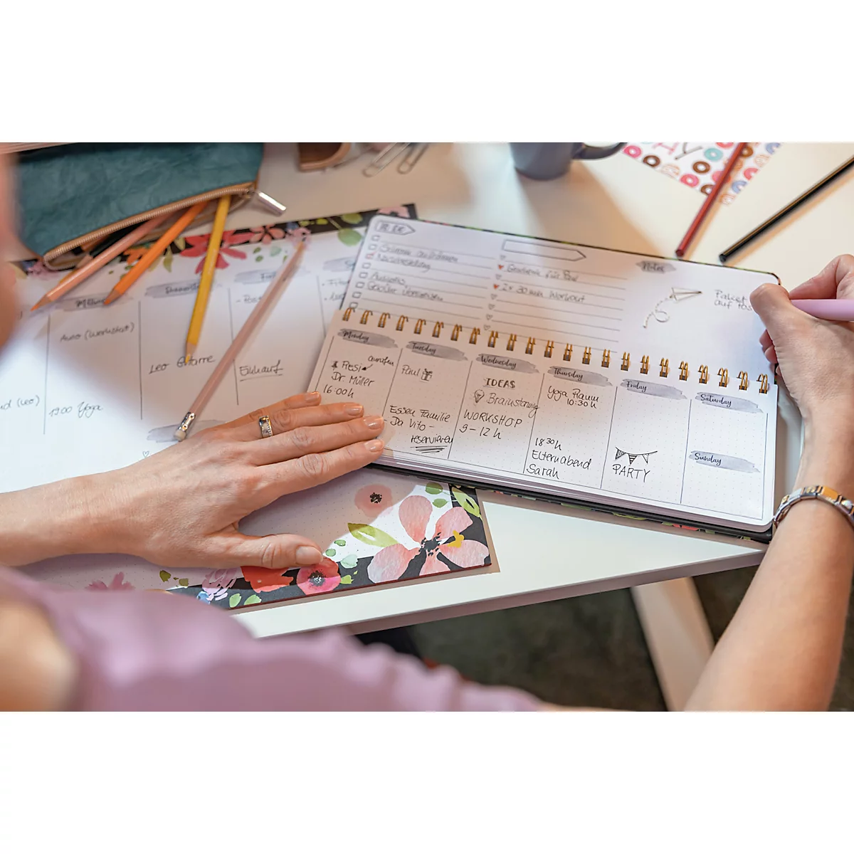Personne écrit dans un agenda hebdomadaire. Stylos, notes et un cahier floral sont sur la table.