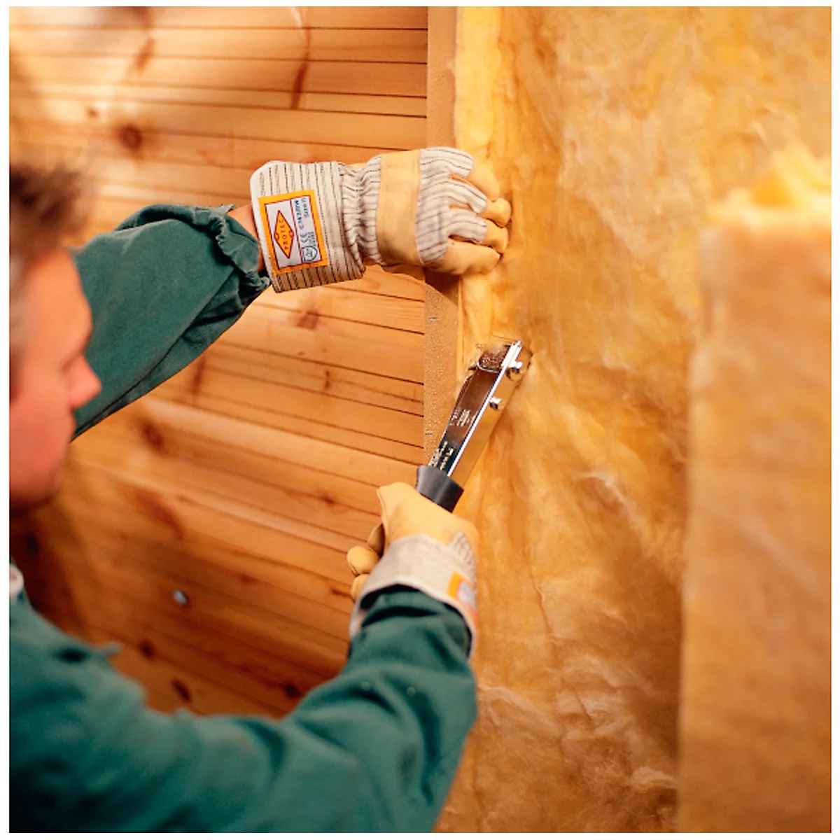 Un bricoleur fixe de l'isolant à une structure en bois avec une agrafeuse. Il porte des gants et une combinaison de travail verte.