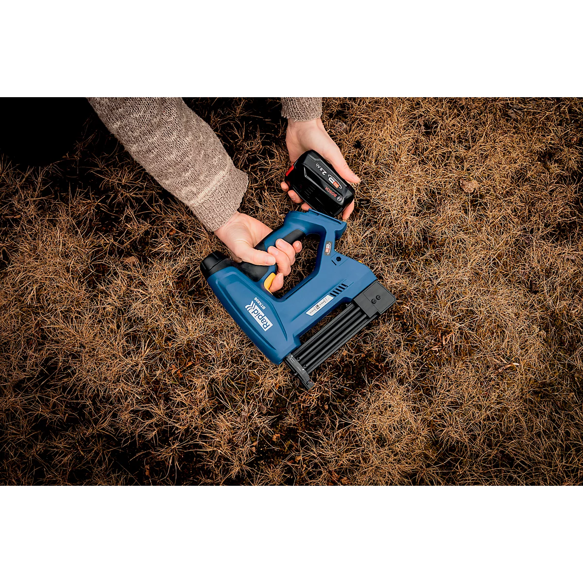 Mains tenant une agrafeuse bleue et une batterie au-dessus d'herbe sèche.
