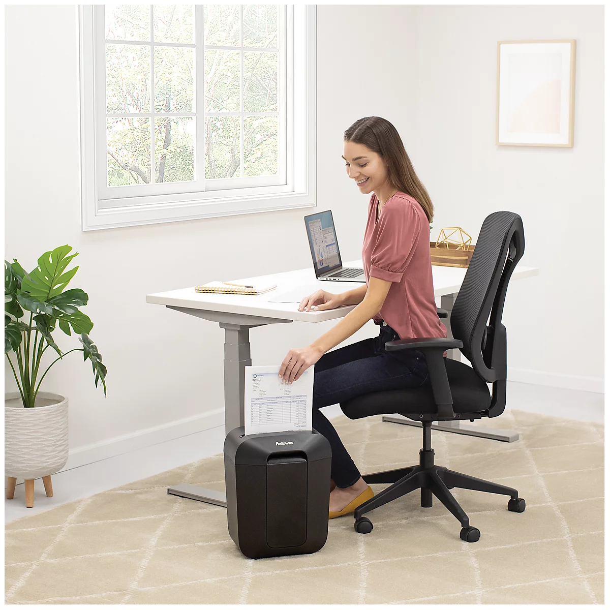 Eine Frau in einem hellen Homeoffice schreddert Dokumente in einem schwarzen Aktenvernichter neben ihrem Schreibtisch.