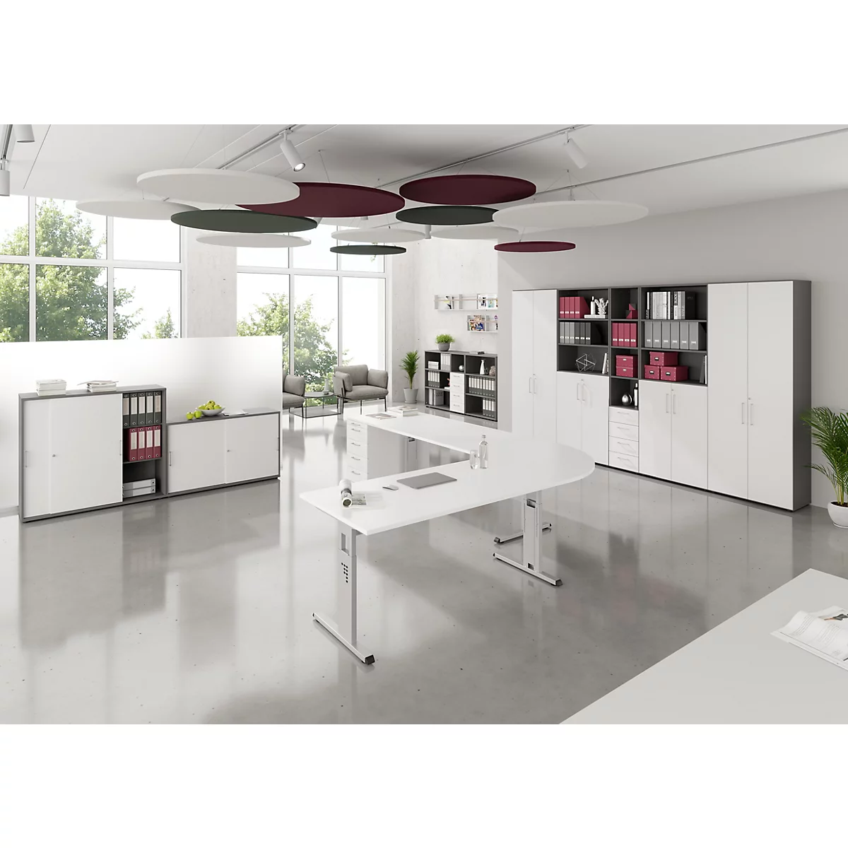 Ein helles Büro mit weißen Schreibtischen, Schränken und runden Deckenplatten. Fenster zeigen einen grünen Ausblick.