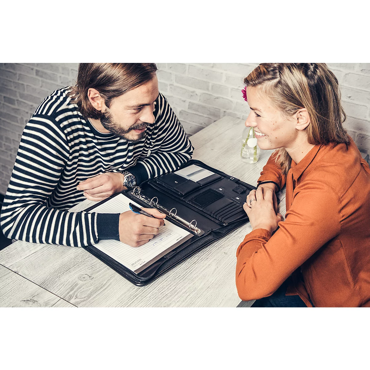 A man in a striped sweater writes in a folder while a woman sits opposite him smiling.