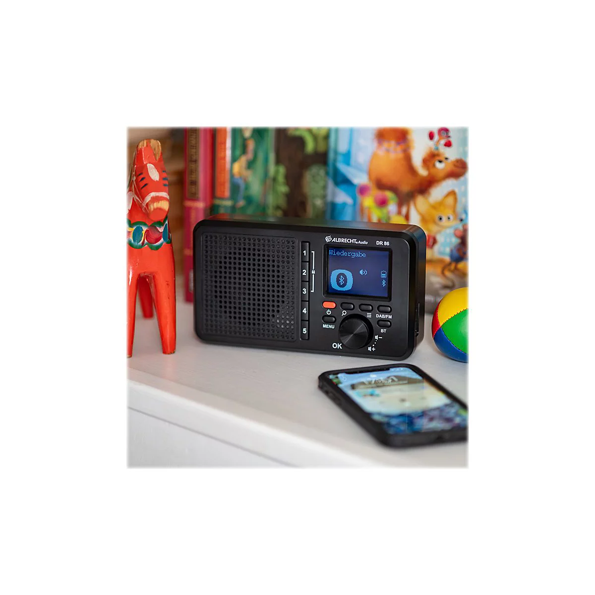 Schwarzes Radio mit blauem Display, daneben ein Smartphone. Auf einem Regal steht ein Holzpferd.
